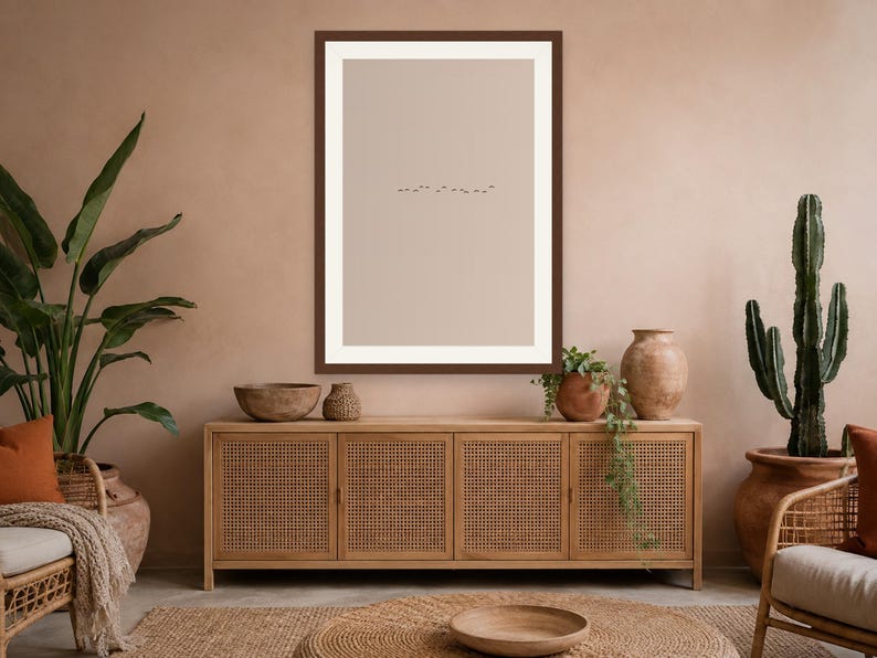 May include: A rectangular wall art print featuring a minimalist beige background with a small, centered line of tiny black birds in flight, enclosed within a dark brown frame, displayed above a wooden sideboard with four cane-paneled doors.