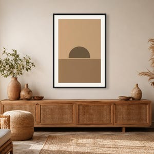 May include: A framed rectangular wall art print featuring a minimalist graphic design of a dark brown semi-circle sun resting on a horizon line, set against a light tan background with a medium brown lower section, enclosed in a black frame.