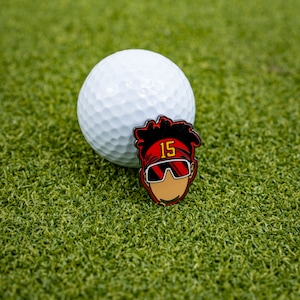 May include: A white golf ball with a red and black enamel pin featuring a cartoon character wearing a red headband with the number "15" on it and sunglasses.
