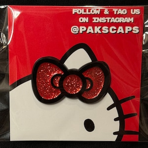 May include: Red glitter Hello Kitty pin with a black outline. The pin features a red glitter bow with a black outline. The text on the card reads "FOLLOW & TAG US ON INSTAGRAM @PAKSCAPS".