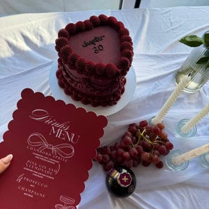 May include: A heart-shaped, burgundy cake with the text "Chapter 30" sits on a white plate. A red menu card with drink options, grapes, and a decorative bottle are also present. The scene is set on a white tablecloth.