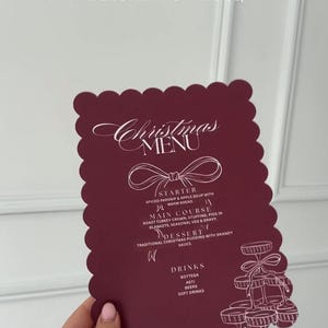 Puede incluir: Una tarjeta de menú navideño burdeos con borde festoneado, con escritura elegante y una ilustración detallada de un soporte de postre en niveles. El menú enumera un entrante, plato principal, postre y bebidas.