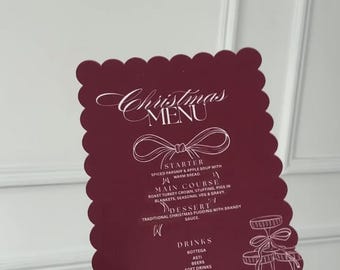 May include: A burgundy Christmas menu card with a scalloped edge, featuring elegant script and a detailed illustration of a tiered dessert stand. The menu lists a starter, main course, dessert, and drinks.