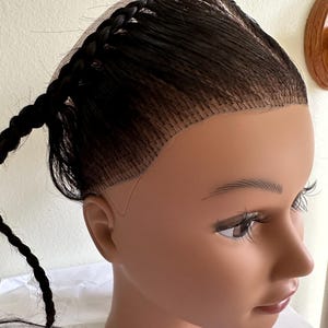 May include: A dark brown wig featuring a sheer lace front base with a visible hairline. The hair is styled with a single braid running along the side of the head, extending into a loose tail. The wig is displayed on a smooth, neutral-toned mannequin head.