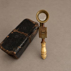 May include: A folding magnifying glass with a circular brass frame, a rectangular brass hinge mechanism, and a cream-coloured turned handle. Beside it sits a rectangular black leather case with visible wear, featuring a hinged lid and embossed details.