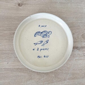 Peut inclure: Une assiette ronde en céramique blanc cassé avec une illustration bleue de pain et de poissons. L'assiette porte l'inscription "5 pains et 2 poissons, Marc 6:41" en bleu. L'assiette est posée sur une surface en bois clair.