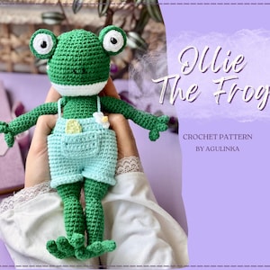 Könnte beinhalten: Eine grüne, gehäkelte Froschpuppe mit großen weißen Augen, die eine hellblaue Latzhose mit kleiner Tasche und einer weißen Blume trägt. Der Text auf der rechten Seite lautet Ollie The Frog, Crochet Pattern by Agulinka, vor violettem Hintergrund.