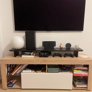 Può includere: Un moderno mobile da intrattenimento con una grande televisione nera montata sopra. Il mobile in legno presenta ripiani aperti con libri, elettronica e un mobile bianco. Un ripiano nero contiene una console di gioco, controller e oggetti decorativi.