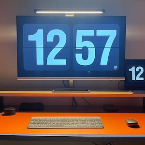 May include: A desk workspace with a computer monitor and laptop displaying the time 12:57. The monitor and laptop are on a wooden shelf above a desk with an orange desk mat, a keyboard, and a mouse.