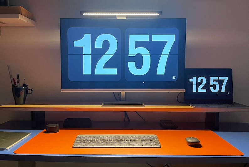 May include: A desk workspace with a computer monitor and laptop displaying the time 12:57. The monitor and laptop are on a wooden shelf above a desk with an orange desk mat, a keyboard, and a mouse.