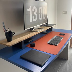 May include: A blue desk with a red desk mat, a black notebook, a keyboard, a mouse, and a black speaker. A wooden shelf above the desk holds a computer monitor with a digital clock displaying 13:00.