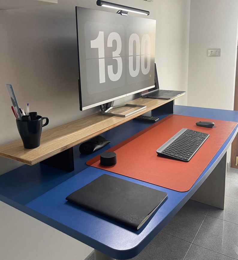 May include: A blue desk with a red desk mat, a black notebook, a keyboard, a mouse, and a black speaker. A wooden shelf above the desk holds a computer monitor with a digital clock displaying 13:00.