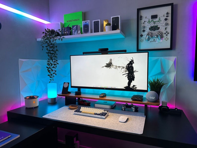 May include: A black desk with a computer monitor, keyboard, mouse, and other accessories. The desk is lit with blue and pink LED lights. There is a plant hanging from the shelf above the desk. The wall behind the desk is decorated with a white geometric pattern and a framed picture of a video game controller.