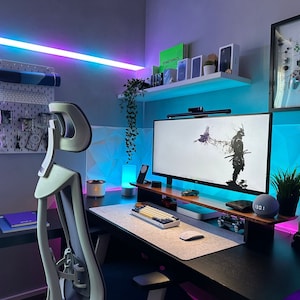 May include: A modern home office setup with a black desk, a white gaming chair, a large monitor displaying a black and white image of a samurai, a keyboard, a mouse, and a smart speaker. The walls are painted a light gray and have blue and purple LED lights.