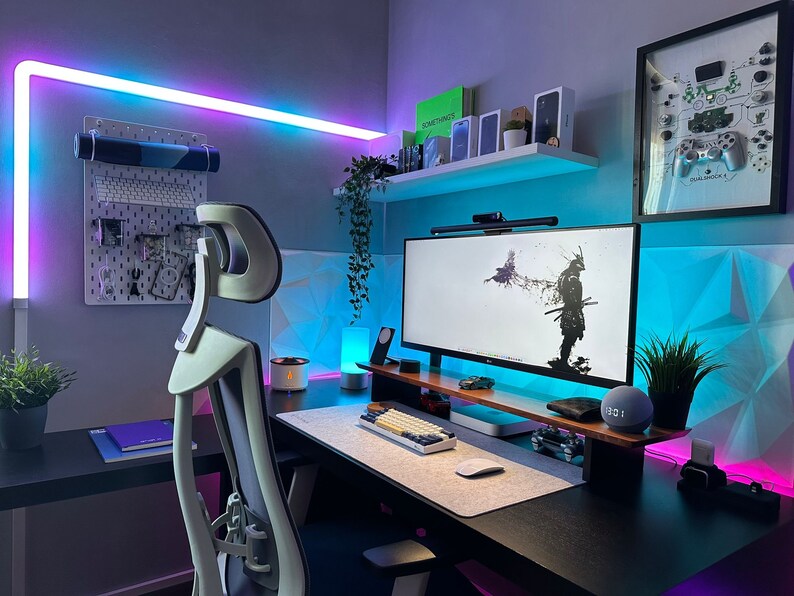 May include: A modern home office setup with a black desk, a white gaming chair, a large monitor displaying a black and white image of a samurai, a keyboard, a mouse, and a smart speaker. The walls are painted a light gray and have blue and purple LED lights.
