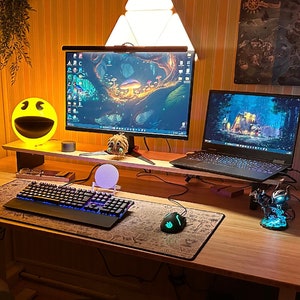 Puede incluir: Un escritorio de madera con un teclado negro, un ratón verde y negro, una figura negra y azul, una lámpara blanca, una lámpara amarilla con forma de cara sonriente, un portátil negro y un monitor negro con una escena de bosque en la pantalla.