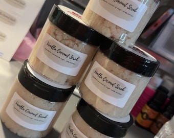 May include: A stack of six clear plastic jars with black lids, each labeled "Vanilla Coconut Scrub." The jars are filled with a light-colored, granular substance. The background includes a stack of clear plastic containers and a sheet of labels.