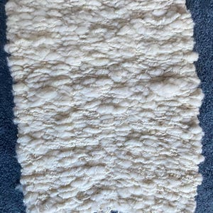 May include: A rectangular, off-white woven rug with a textured, nubby surface. The rug has a fringe detail along the edges and is set against a blue background. The weave appears to be thick and dense, suggesting a handmade or artisanal quality.