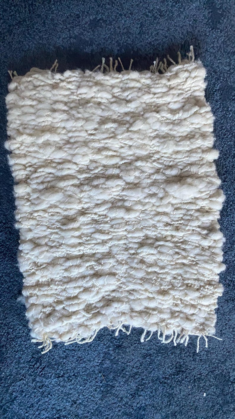 May include: A rectangular, off-white woven rug with a textured, nubby surface. The rug has a fringe detail along the edges and is set against a blue background. The weave appears to be thick and dense, suggesting a handmade or artisanal quality.