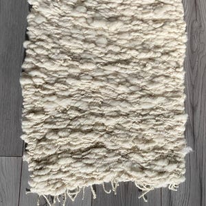May include: Cream-colored, hand-woven rug with a textured, chunky weave. The rectangular rug has a fringe detail along one edge. The rug is made of a soft, natural fiber and is suitable for a variety of home decor styles.
