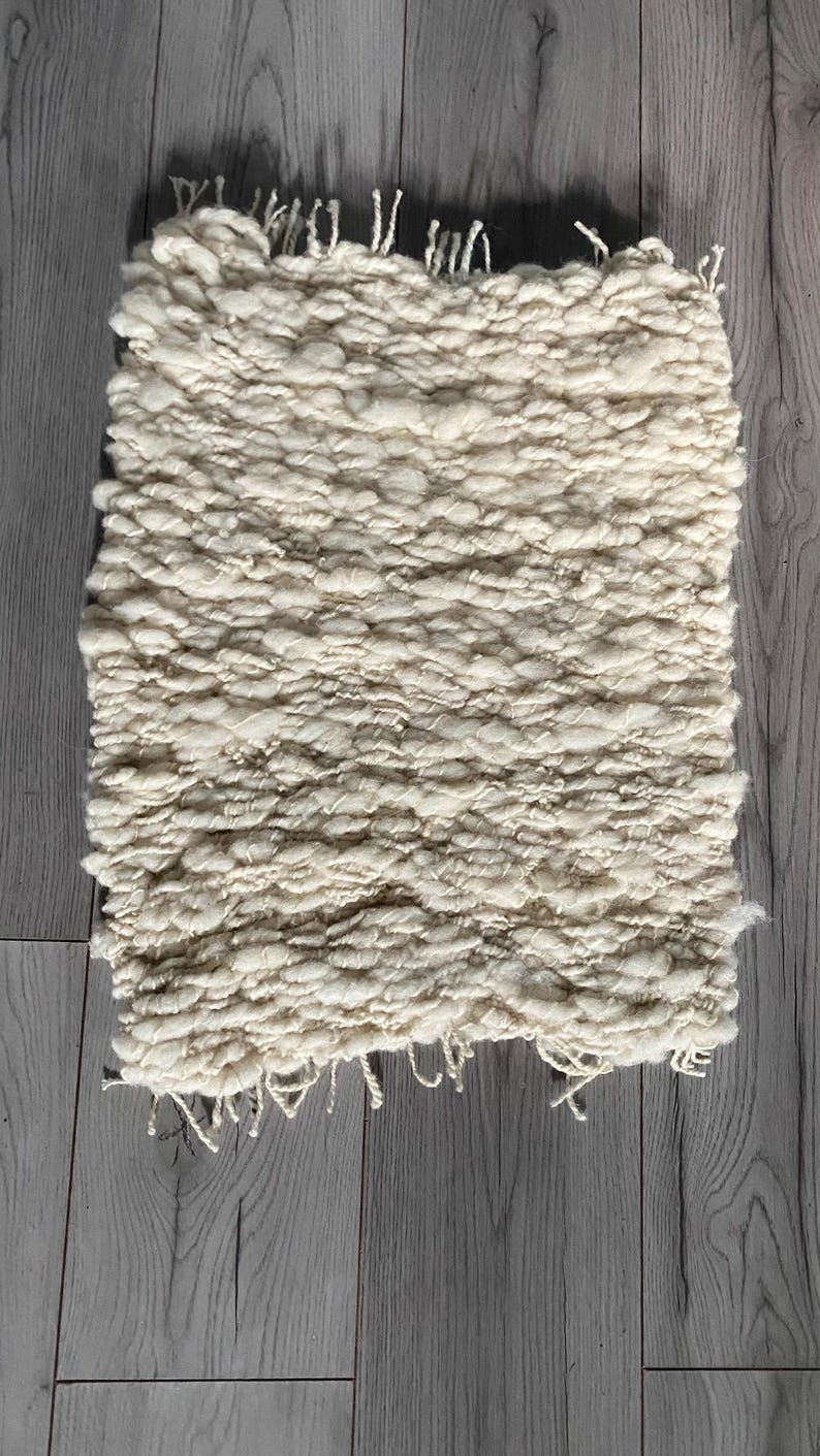 May include: Cream-colored, hand-woven rug with a textured, chunky weave. The rectangular rug has a fringe detail along one edge. The rug is made of a soft, natural fiber and is suitable for a variety of home decor styles.