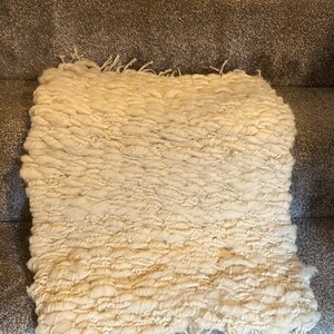 May include: Cream-colored, hand-woven rug with a textured, looped design. The rug has a rectangular shape and a fringe along one edge. The thick, soft material suggests a cozy, comfortable feel. The rug is placed on a gray carpeted surface.