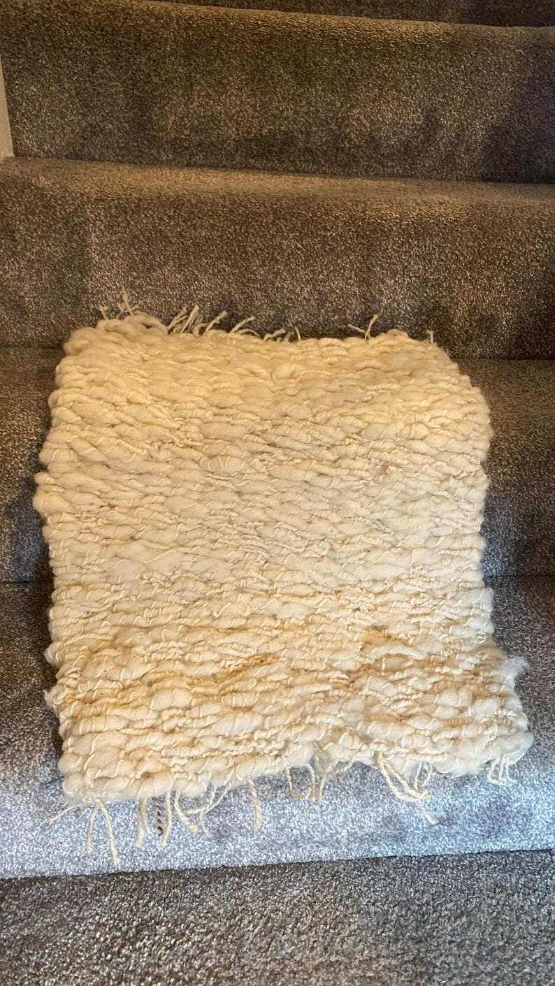 May include: Cream-colored, hand-woven rug with a textured, looped design. The rug has a rectangular shape and a fringe along one edge. The thick, soft material suggests a cozy, comfortable feel. The rug is placed on a gray carpeted surface.