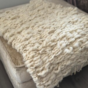 May include: A cream-coloured, chunky knit throw blanket. The blanket has a thick, textured appearance with visible loops and a soft aesthetic. The blanket is draped over a neutral-toned surface, suggesting a cosy home decor item.