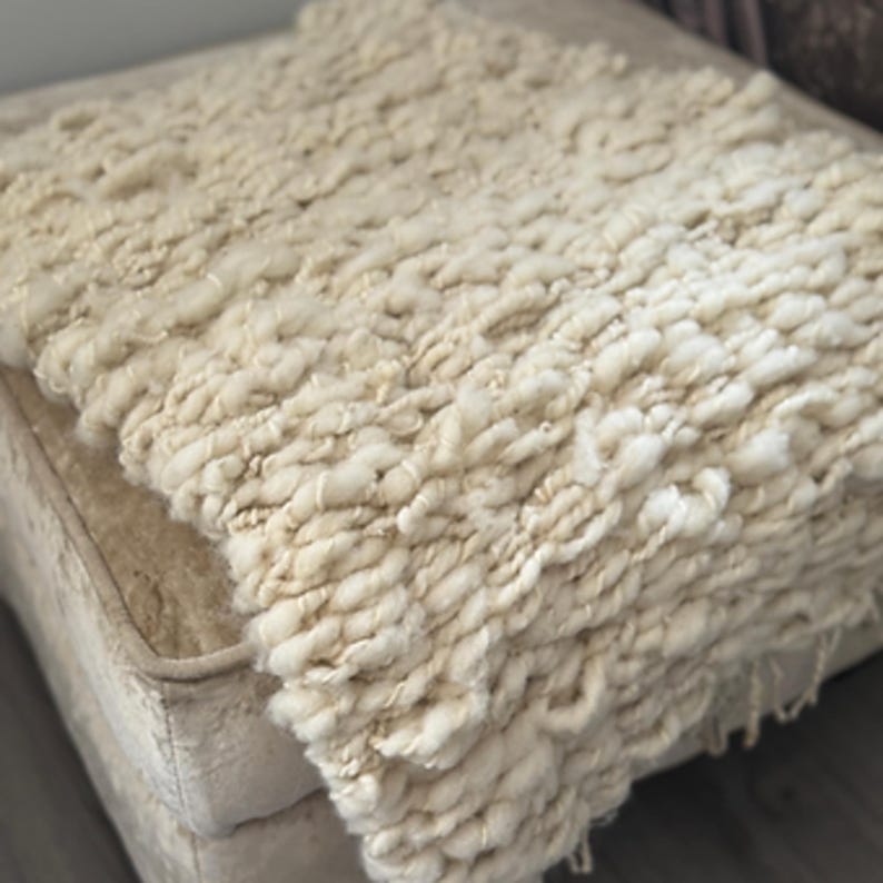 May include: Cream-colored, chunky knit throw blanket. The blanket has a thick, textured appearance with visible loops and a soft, inviting aesthetic. The blanket is draped over a neutral-toned surface, suggesting a cozy home decor item.
