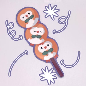 May include: A sticker featuring three cartoon owls on a stick, resembling a Japanese dango skewer. The owls are orange and white with black eyes and green leaves in their beaks. The background is a light purple.