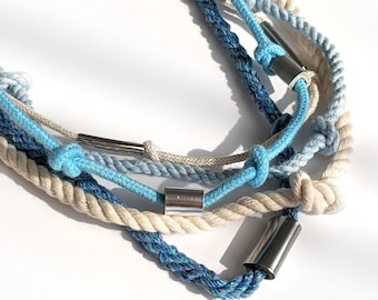 May include: A multi-strand necklace with a nautical theme. The necklace is made of thick, twisted rope in shades of blue and cream, with silver-toned metal accents. Knots and metal tubes add detail to the necklace.