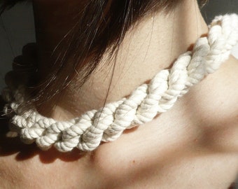 May include: A cream-coloured, braided rope necklace. The necklace is thick and chunky, with a textured appearance. The rope is tightly woven, creating a bold accessory. The necklace is worn around the neck.
