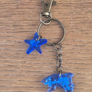 Portachiavi a forma di squalo fatto a mano con plastica oceanica riciclata e resina epossidica. Ogni vendita finanzia la pulizia dell'oceano. L'articolo viene consegnato in una scatola regalo di cartone.