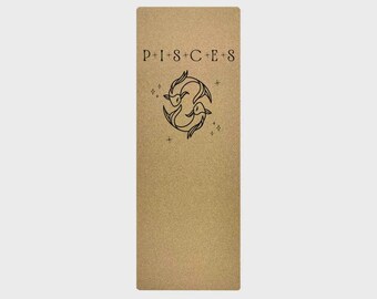 May include: A rectangular yoga mat with a textured, tan surface. The mat features the word "PISCES" in black capital letters at the top, above a black illustration of two fish. Small star-like designs are also present.