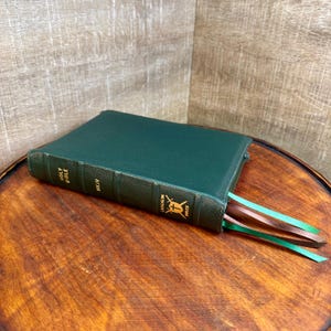 May include: A dark green hardcover Bible with a textured spine featuring gold embossed text that reads HOLY BIBLE, NKJV, and LAUGHLIN BIBLES with a shield and sword emblem. Three ribbon bookmarks in green and brown extend from the bottom edge.