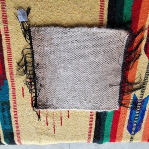 May include: A close-up of a small, white woven rug with black fringe. The rug is on top of a larger, colourful rug with a striped pattern.