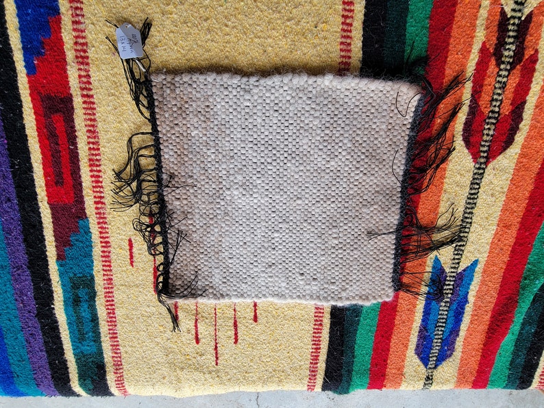 May include: A close-up of a small, white woven rug with black fringe. The rug is on top of a larger, colourful rug with a striped pattern.