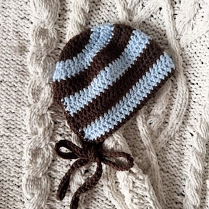 May include: A crocheted bonnet featuring alternating horizontal stripes in dark brown and light blue yarn. The headwear includes two dark brown crocheted ties attached at the base, designed to be fastened under the chin for a secure fit.
