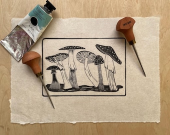 May include: A black and white linocut print of five mushrooms with white spots on their caps. The print is on a beige paper background. Two wood carving tools are also in the image.