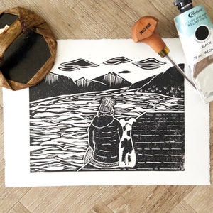 Può includere: Stampa a linoleum in bianco e nero di una persona con un cappello e un cane seduti su un bordo che si affaccia su uno specchio d'acqua con montagne sullo sfondo. La stampa è su carta bianca e nell'immagine sono presenti uno strumento da intaglio e un tubo di inchiostro da rilievo nero.