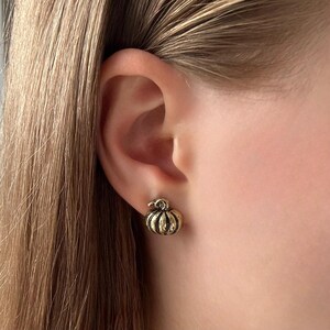 May include: A gold-coloured pumpkin stud earring. The earring is worn on the ear of a person. The pumpkin has detailed ridges and a small stem. The earring is a fall-themed accessory.