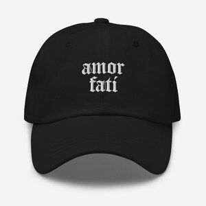 May include: Black baseball cap with the words "amor fati" embroidered in white gothic font across the front. The cap has a curved brim and an adjustable strap at the back. A classic, casual accessory.