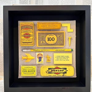 Puede incluir: Una exhibición enmarcada con artículos amarillos vintage. Incluye una caja de cigarrillos, dinero de Monopoly, un bolígrafo, un avión de juguete, un sello, una caja de cerillas y un kit de reparación Dunlop. Los artículos están dispuestos sobre un fondo de tela beige.