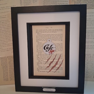 May include: A framed page from the book "Cujo" by Stephen King. The page features a close-up of a bloody handprint on the page, with the text "Cujo" in a circle on the page. The text on the page reads "Mommy, I have to go get the bat..."