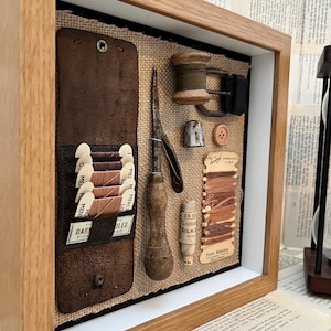 Puede incluir: Una caja de sombra enmarcada con nociones de costura vintage. Incluye un portahilos de cuero con carretes, una aguja, un dedal, un botón, una bobina de hilo y una tarjeta de seda trenzada. El marco es de madera clara.