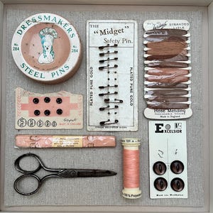 May include: A selection of vintage haberdashery items, including a tin of steel pins, safety pins, thread, buttons, and scissors. The items are displayed on a neutral fabric background, with a variety of colours and textures.