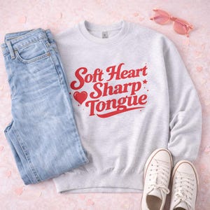 May include: A light gray sweatshirt with the text "Soft Heart Sharp Tongue" in red, paired with light wash jeans, white sneakers, and pink sunglasses. The items are arranged on a pink surface.
