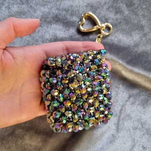 Puede incluir: Un pequeño estuche cuadrado para accesorios cubierto de cuentas iridiscentes multicolores. Tiene un cierre dorado en forma de corazón y un llavero a juego. El estuche se sostiene en una mano sobre un fondo gris.