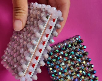 May include: Two beaded card holders. One is light gray with a red stripe, and the other is iridescent with a mix of colors. The beads are small and faceted, giving them a sparkling appearance. The background is a solid pink.