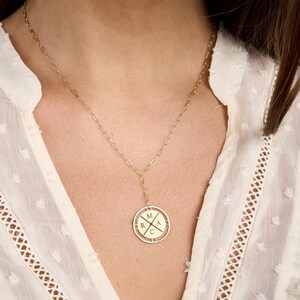 May include: Gold-toned necklace with a delicate chain and a circular pendant. The pendant features the letters M, A, R, and C, arranged in a cross pattern. The necklace is worn against a white blouse.